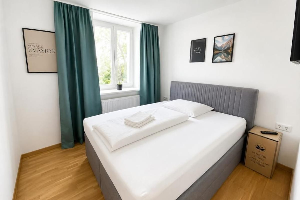 A neatly made bed with white linens and a grey headboard in a room with wooden flooring, a window with curtains, and two framed pictures on the wall.