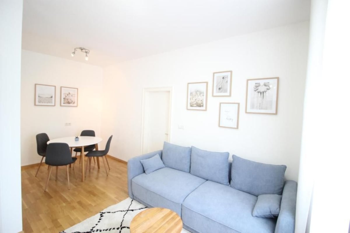 Een moderne woonkamer met een lichtblauwe bank, een ronde houten salontafel, en een eethoek met een witte tafel en zwarte stoelen.