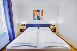 Een tweepersoonsbed met witte lakens, twee nachtkastjes met lampen, een blauw hoofdeinde en een kleurrijk abstract schilderij aan de muur.