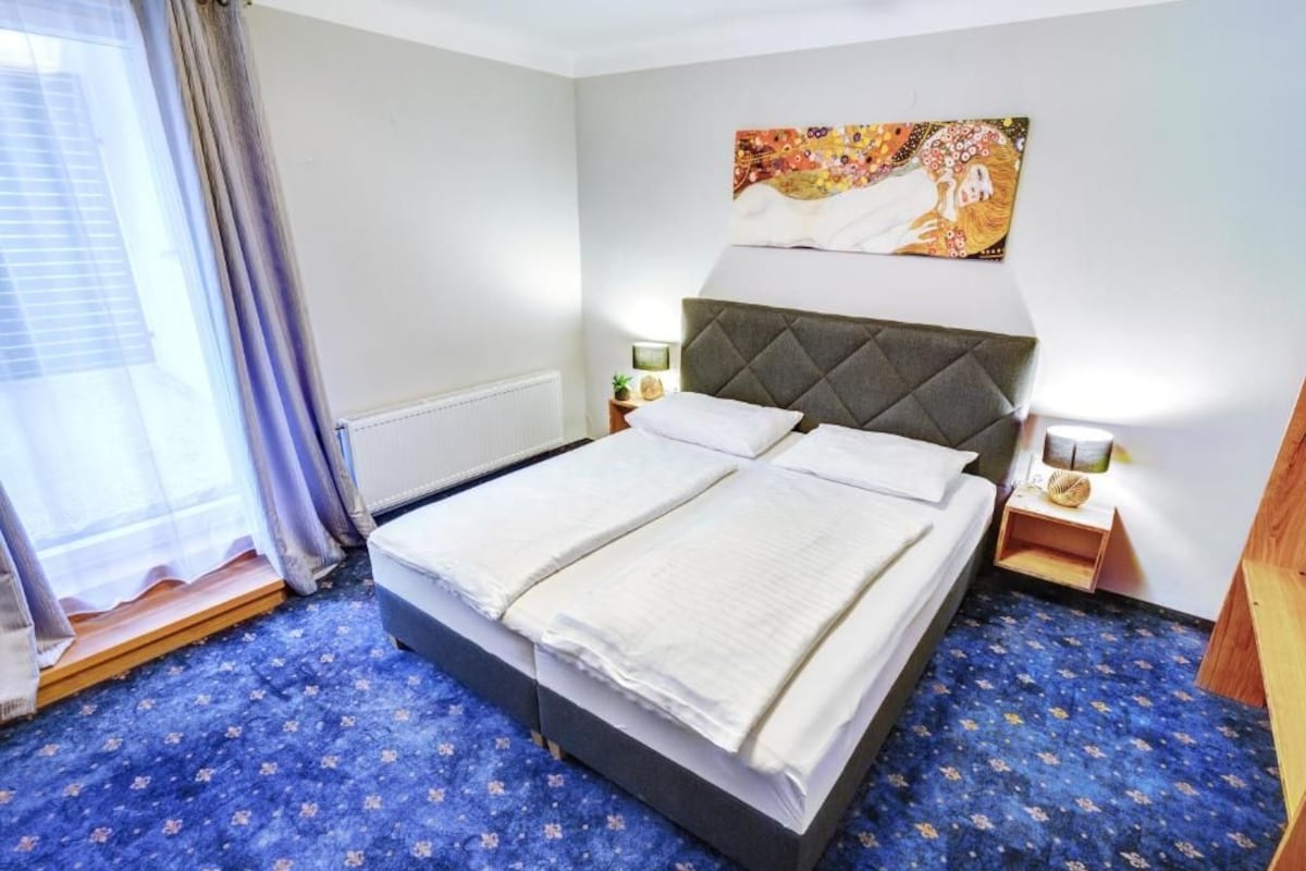 Une chambre d’hôtel avec un lit, une table de chevet, une lampe, un tableau au mur et une fenêtre avec des rideaux.