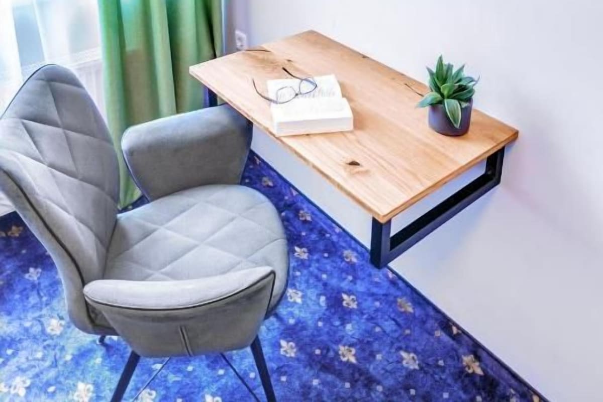Une pièce comprenant un téléviseur fixé au mur, un bureau en bois, une chaise, une plante en pot et une fenêtre avec des rideaux verts.