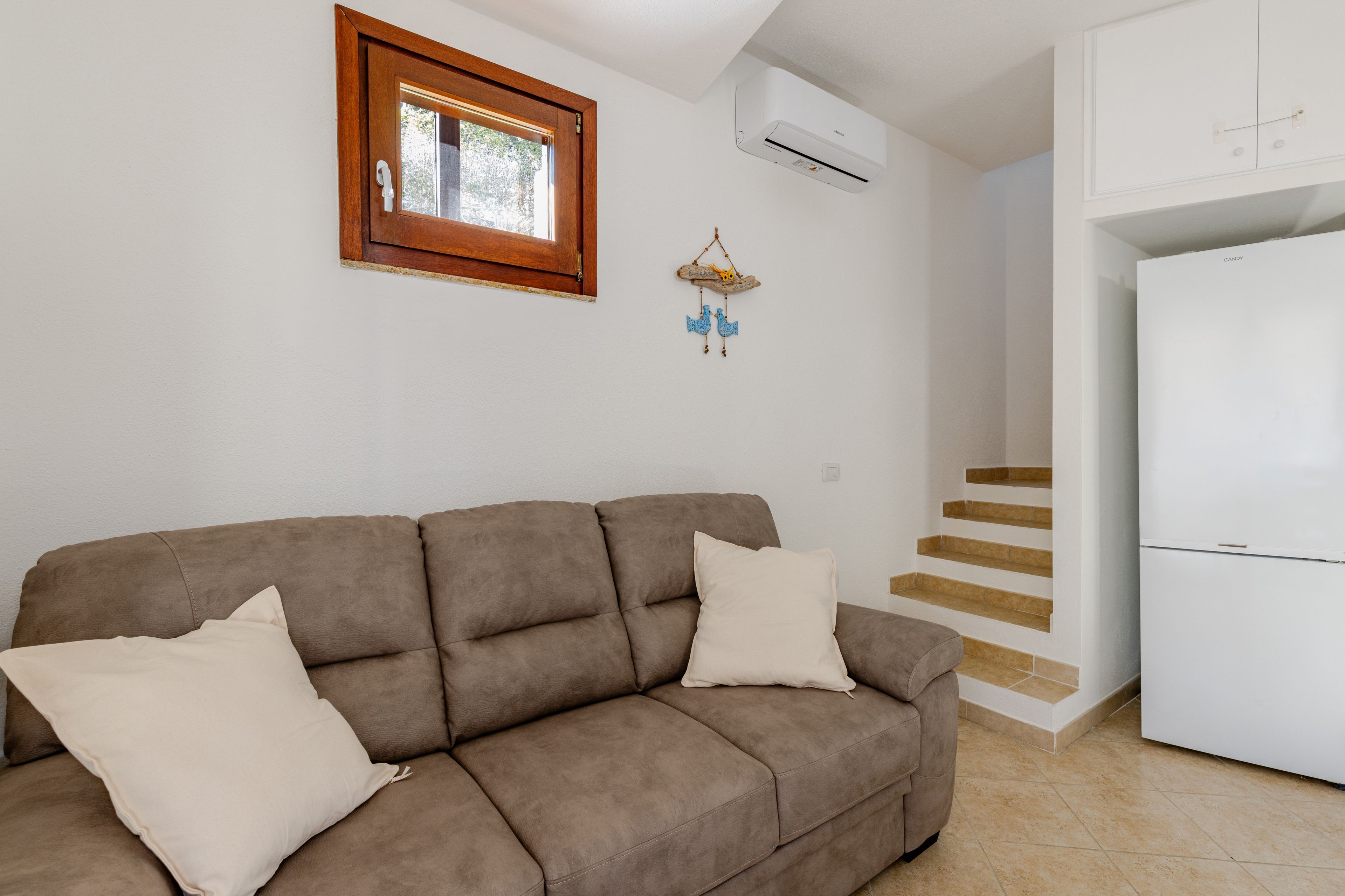 Ein Wohnzimmer mit einer Couch, einem Fenster, einer Klimaanlage, einem Wanddekor und einer Treppe, die in einen Küchenbereich führt.