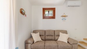 Una sala de estar con un sofá color beige, dos cojines, un ventanal pequeño y un aire acondicionado de pared.