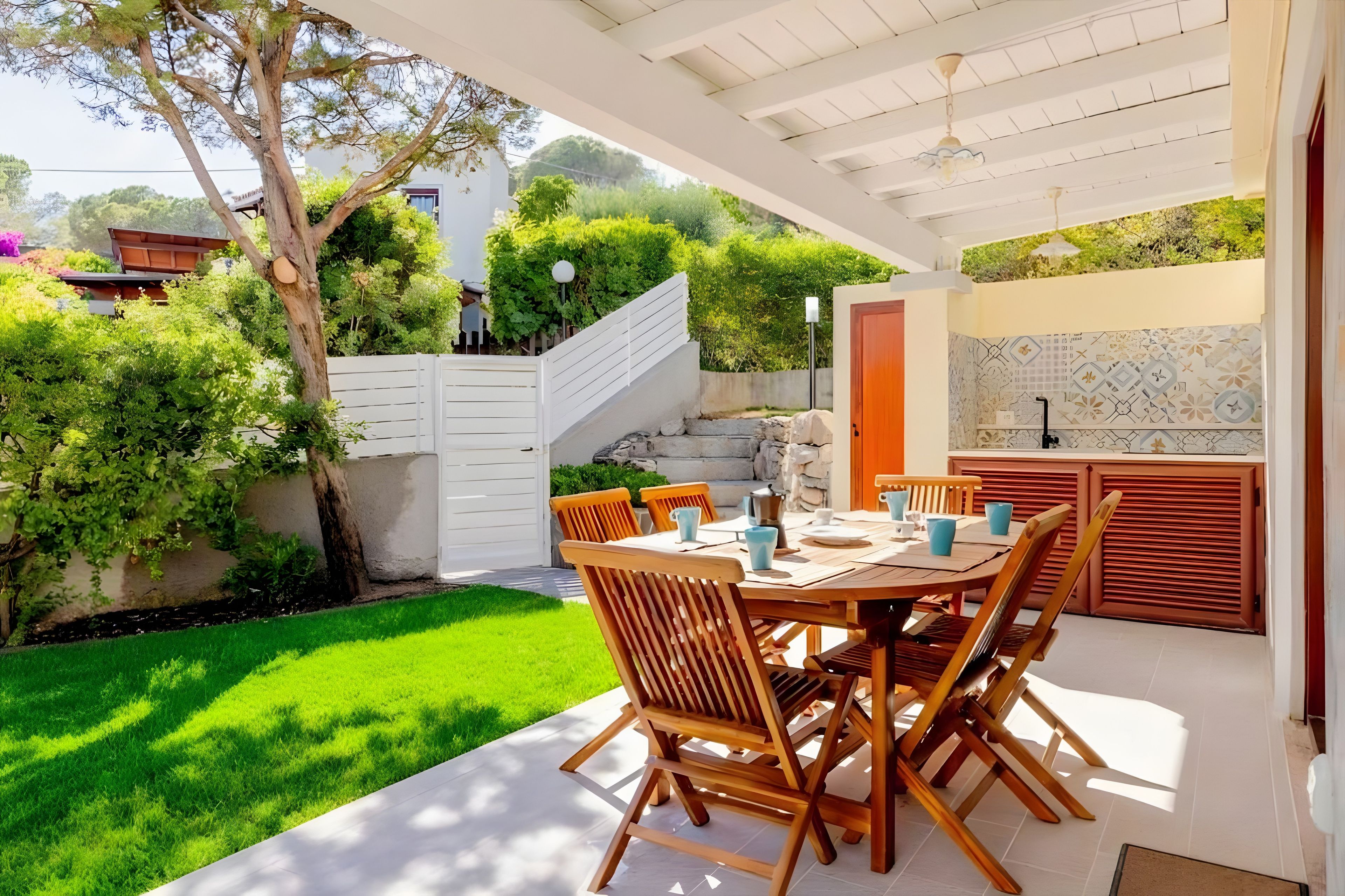 Un patio al aire libre con una mesa lista para comer, rodeado de follaje y una cerca blanca.