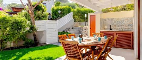 Un patio al aire libre con una mesa lista para comer, rodeado de follaje y una cerca blanca.