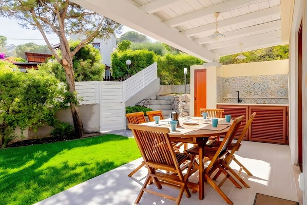 An outdoor patio with a table set for dining, surrounded by greenery and a white fence.