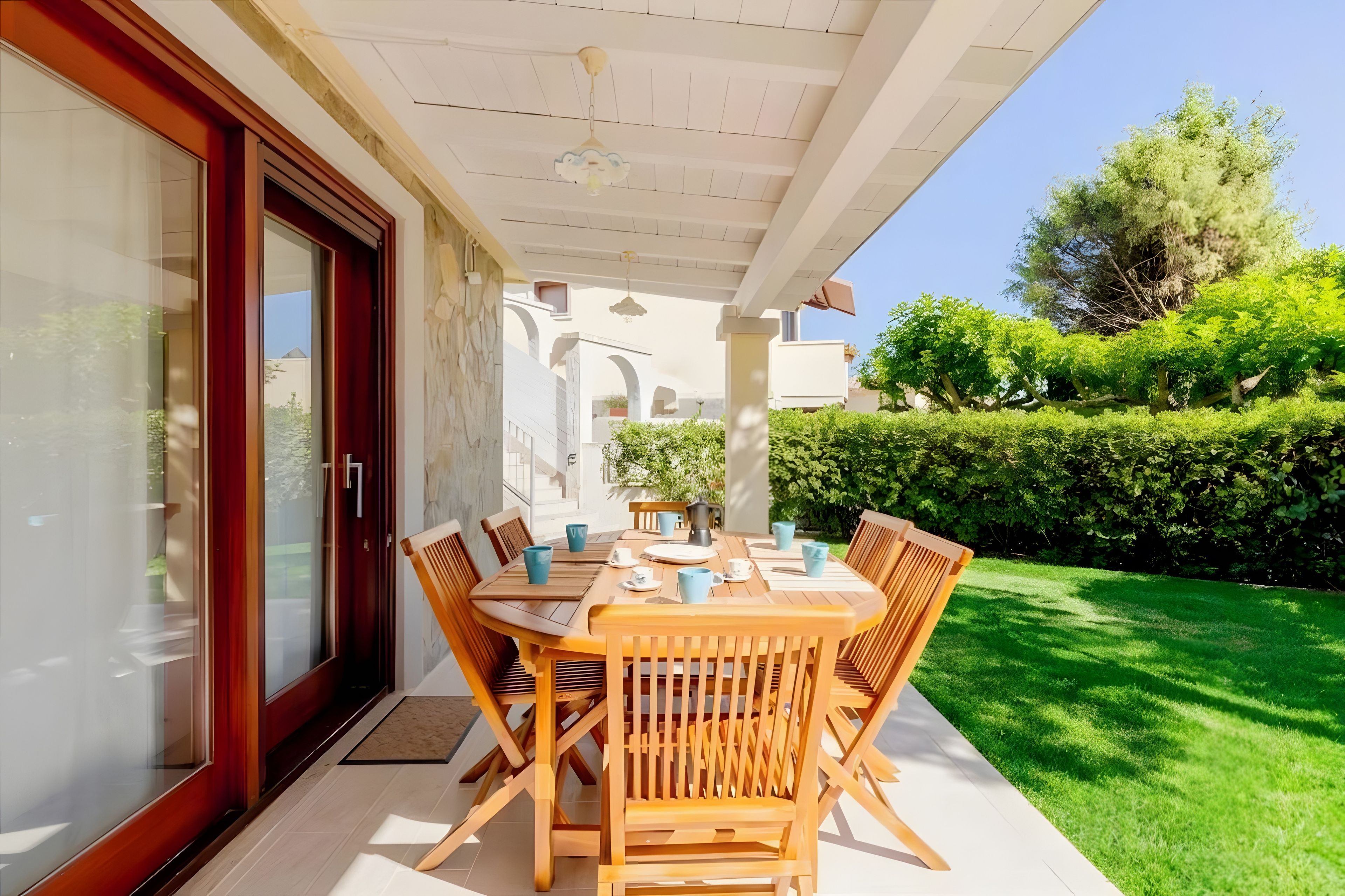 Un porche cubierto con una mesa y sillas de madera, una puerta de vidrio y vistas a un jardín verde.