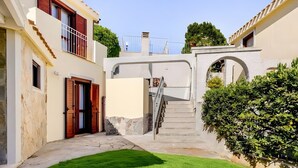 Un patio estrecho con una escalera de piedra que conduce a un balcón en un segundo piso, rodeado de edificios con muros de piedra y un césped bien cuidado.