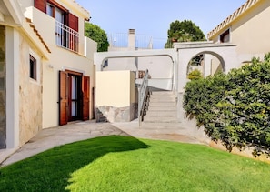 A narrow courtyard with a stone staircase leading to a second-floor balcony, surrounded by buildings with stone walls and a well-maintained lawn. - Villa Le Pavoncelle Home Holiday – Private Garden, 3 Bedrooms, Near the Beach (Eden Rock)