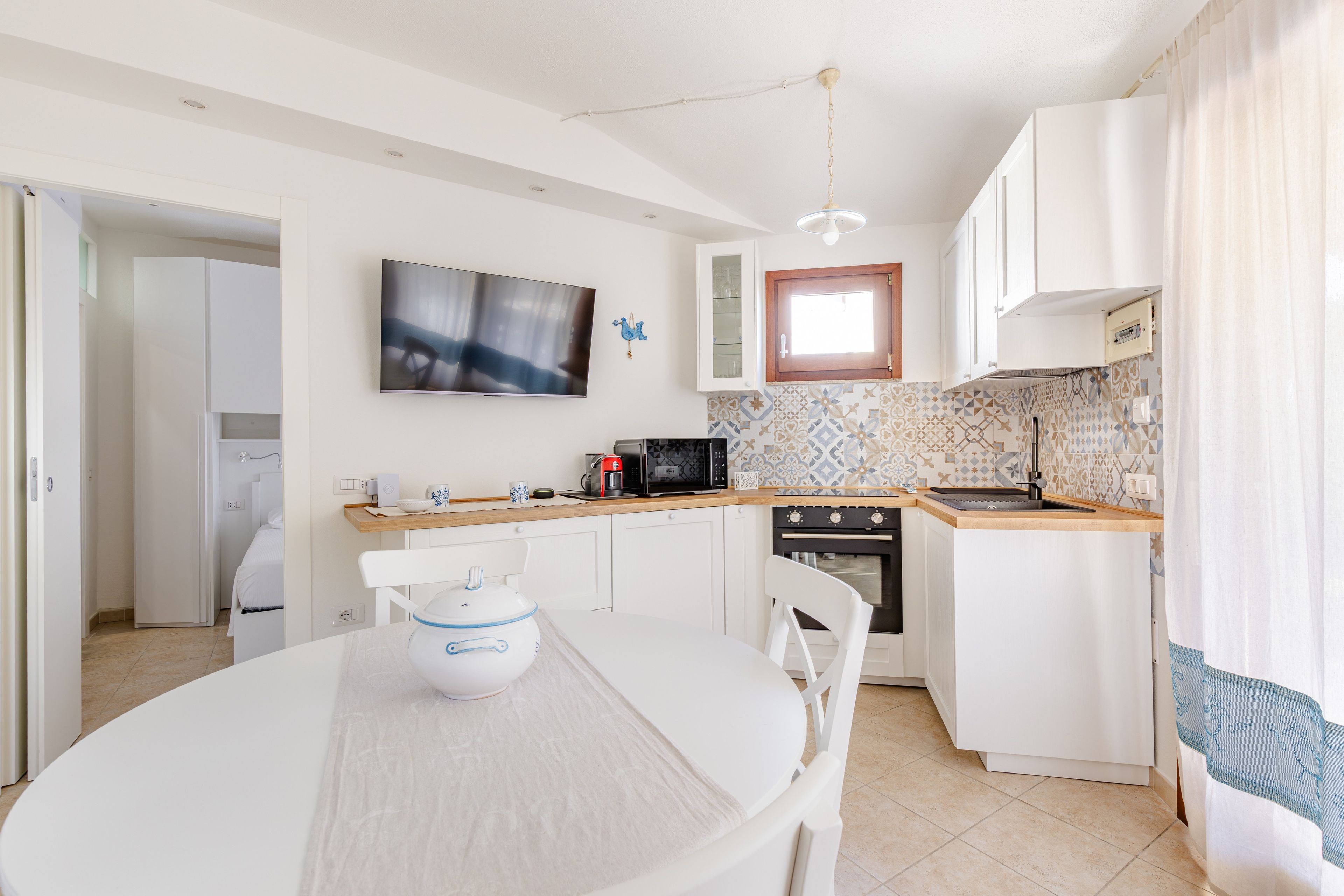 A modern kitchen with white cabinetry, a flat-screen TV, a coffee machine, and a dining area with a table and chairs.