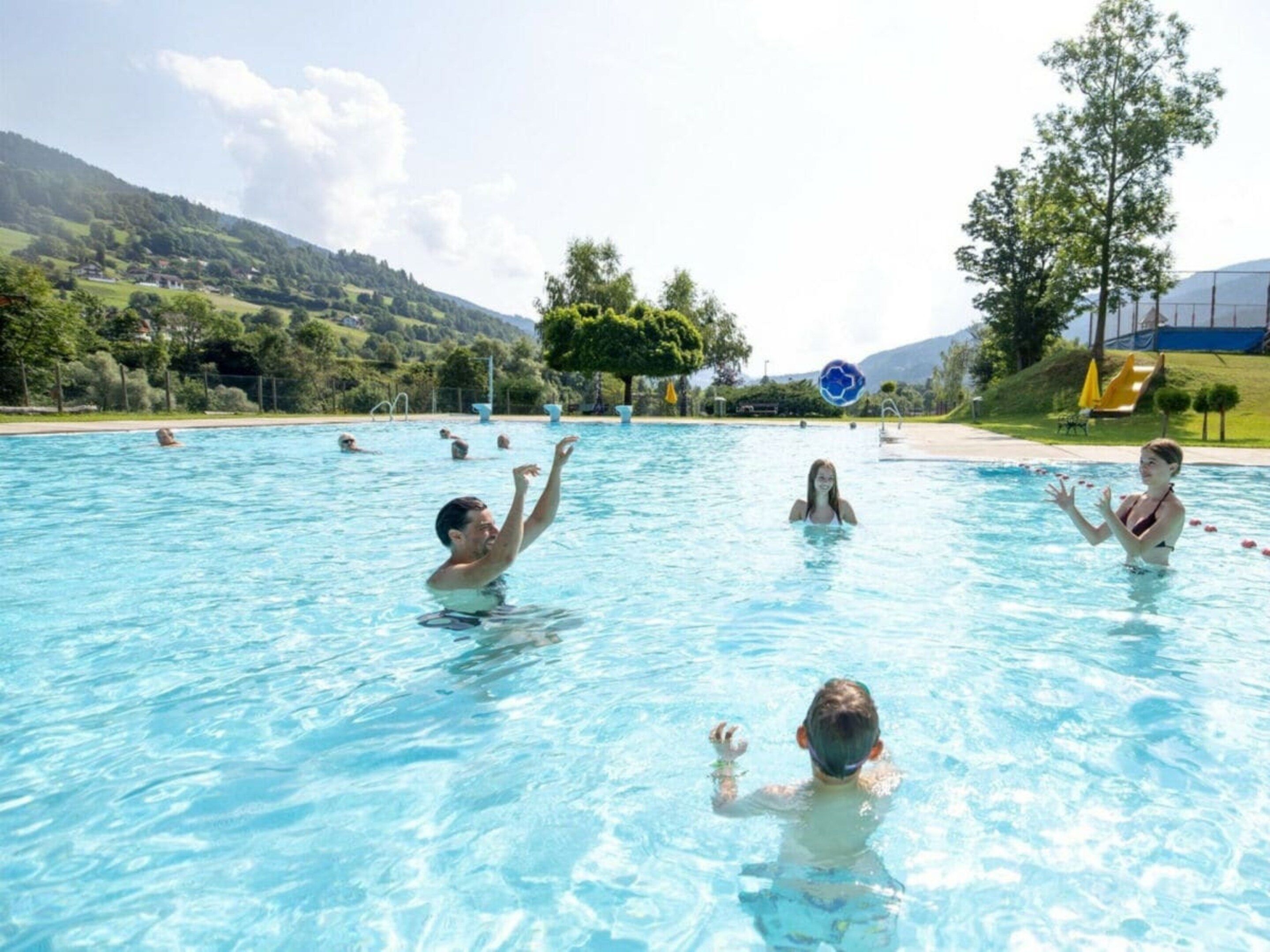 Una piscina con varias personas nadando, rodeada por un paisaje con árboles y colinas.
