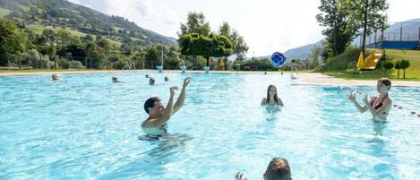 Una piscina con varias personas nadando, rodeada por un paisaje con árboles y colinas.