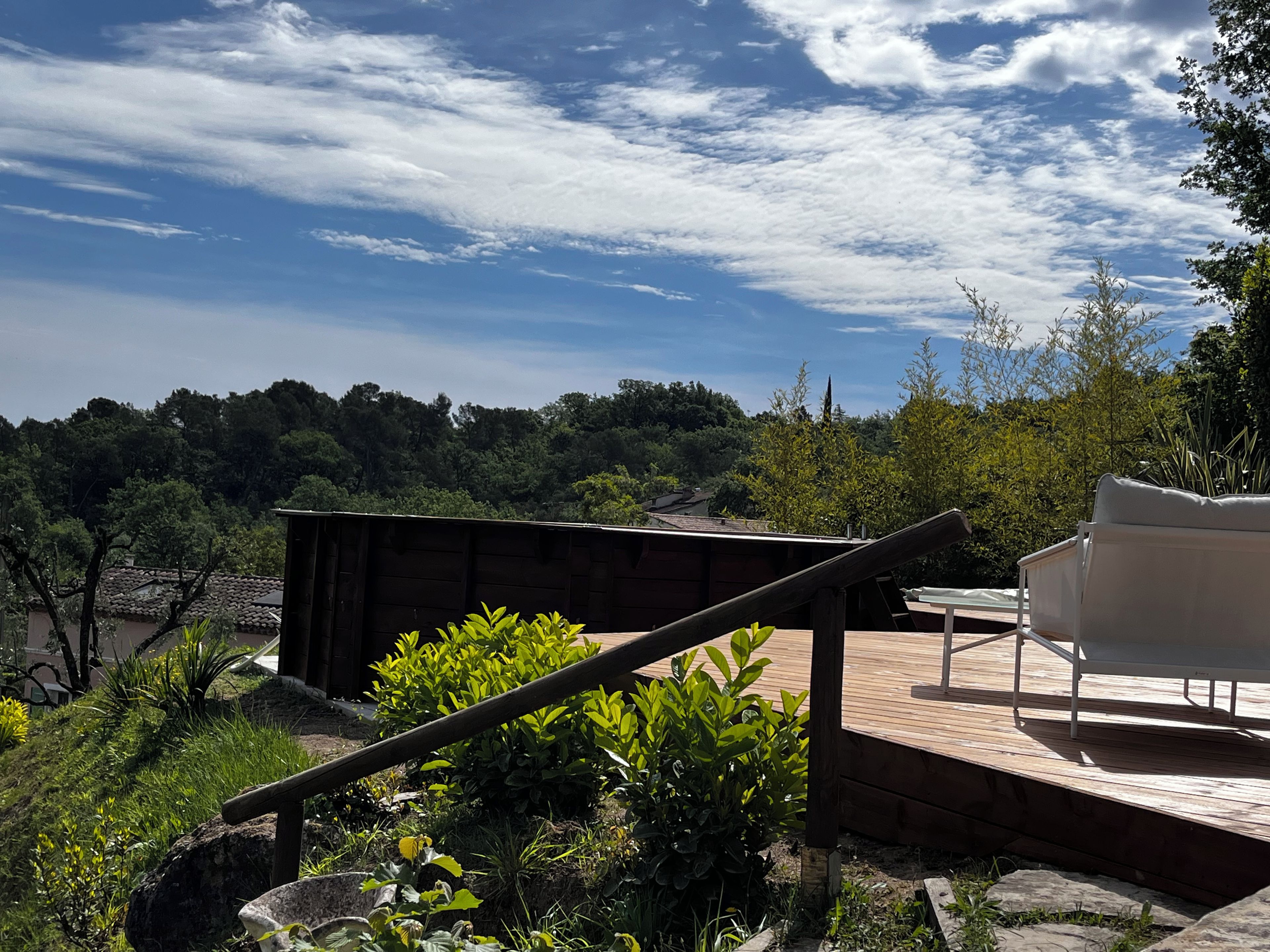 A wooden deck with a white chair, surrounded by greenery and overlooking a forested area.