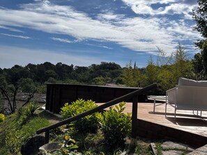 A wooden deck with a white chair, surrounded by greenery and overlooking a forested area.