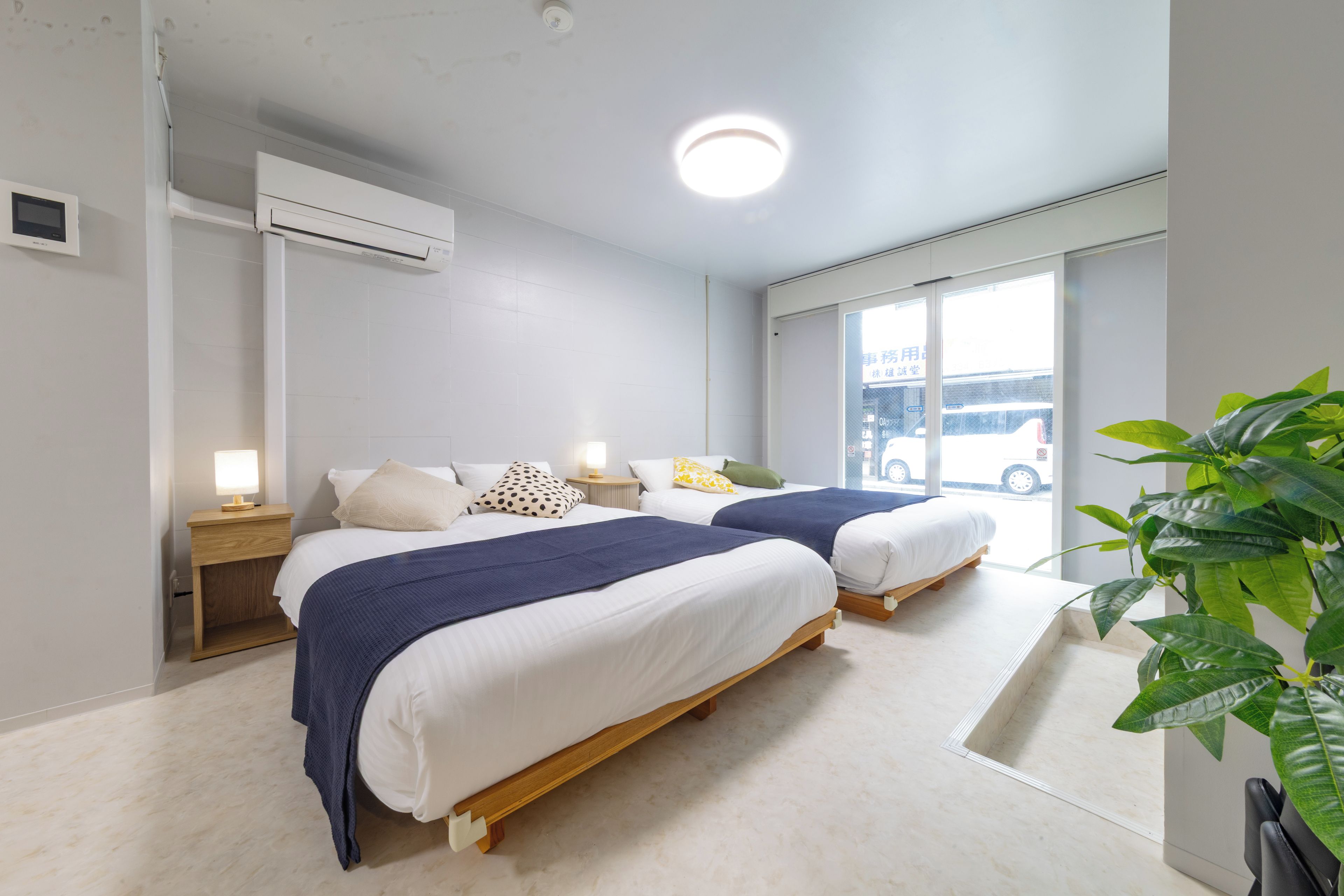 A hotel room with two beds, a bedside table with a lamp, a large window, and a ceiling light.