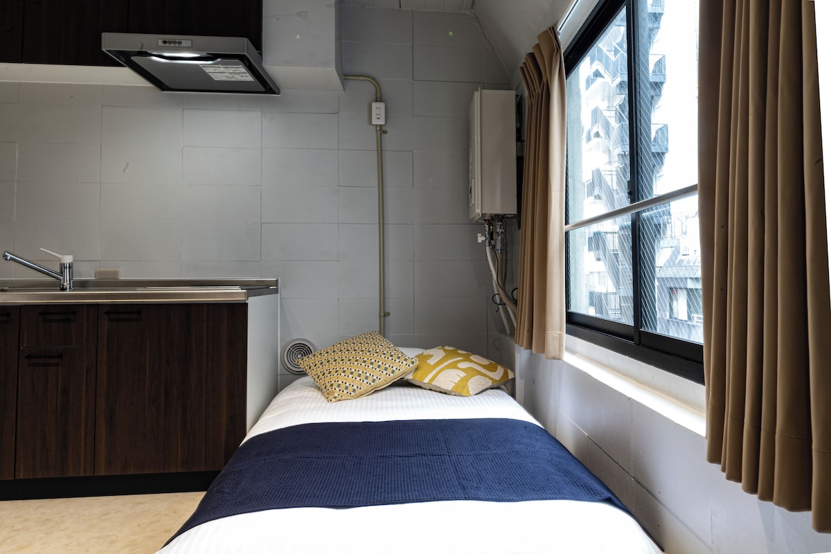 Ein kleines Hotelzimmer mit Küchenzeile, einem Bett mit gemusterten Kissen, einem Fenster mit Vorhängen und Blick auf die Stadtlandschaft.