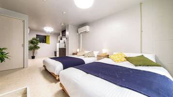 A hotel room with two beds, a small kitchenette, a potted plant, and a window with curtains.