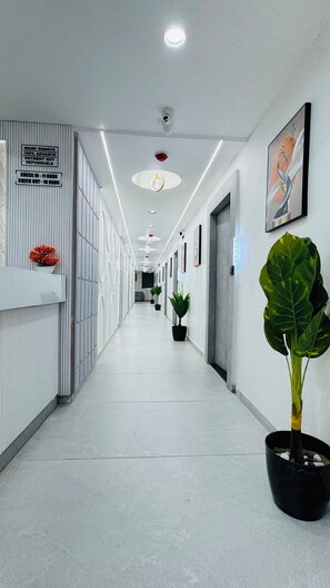 Un pasillo largo y estrecho con un cartel de recepción, un aviso sobre la retirada de mascarillas y una planta a la derecha.