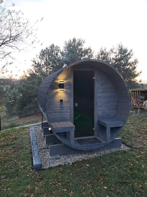 Una estructura cilíndrica de madera con una puerta, escalones que conducen a un banco, y un pequeño espacio exterior con una mesa y leña.