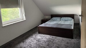 Et soveværelse med skråvæg, en seng med et mønstret sengetæppe, et natbord med en lampe og et vindue med persienner.