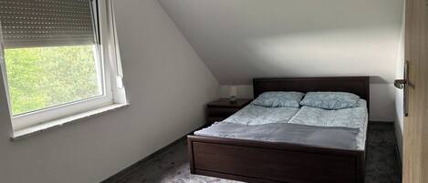 A bedroom with a sloped ceiling, a bed with a patterned bedspread, a nightstand with a lamp, and a window with blinds.