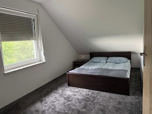 Una camera da letto con soffitto spiovente, un letto con copriletto decorato, un comodino con una lampada e una finestra con persiane.