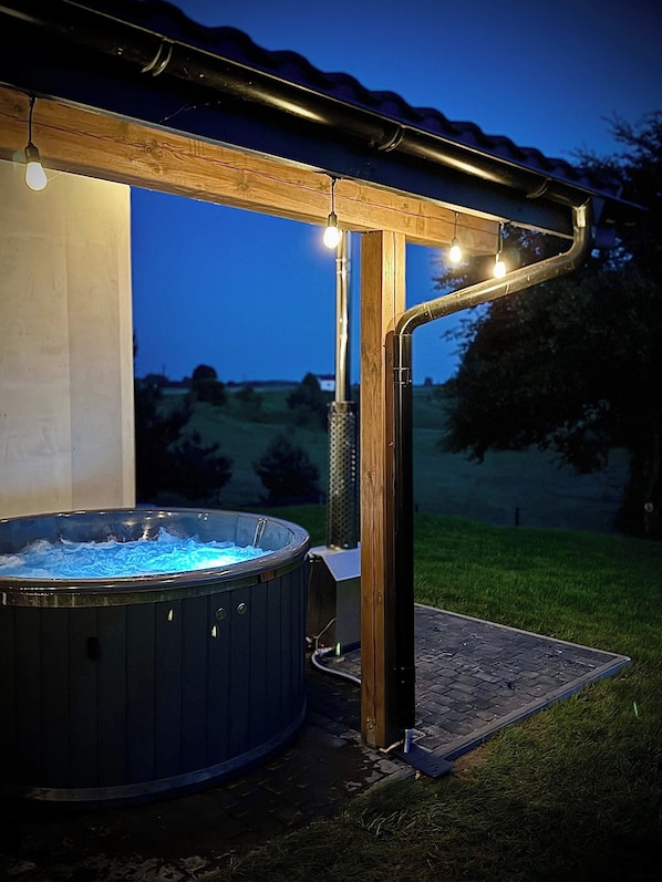 A hot tub under a wooden pergola with string lights, set on a paved patio with a grassy area in the background.