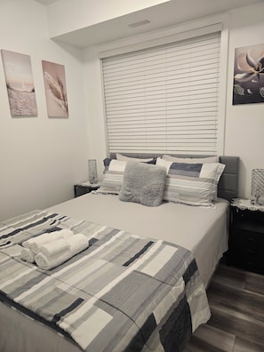 Una cama bien hecha con colcha a rayas y dos almohadas, una mesita de noche con una lámpara y una ventana con persianas.
