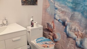 Ett badrum med toalett, handfat och dekorativa element, inklusive en matta med ett snäckmönster och en duschdraperi med ett motiv från en strand.