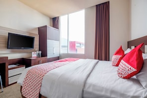 A hotel room with a bed, a television mounted on the wall, a desk with drawers, and a window with curtains.