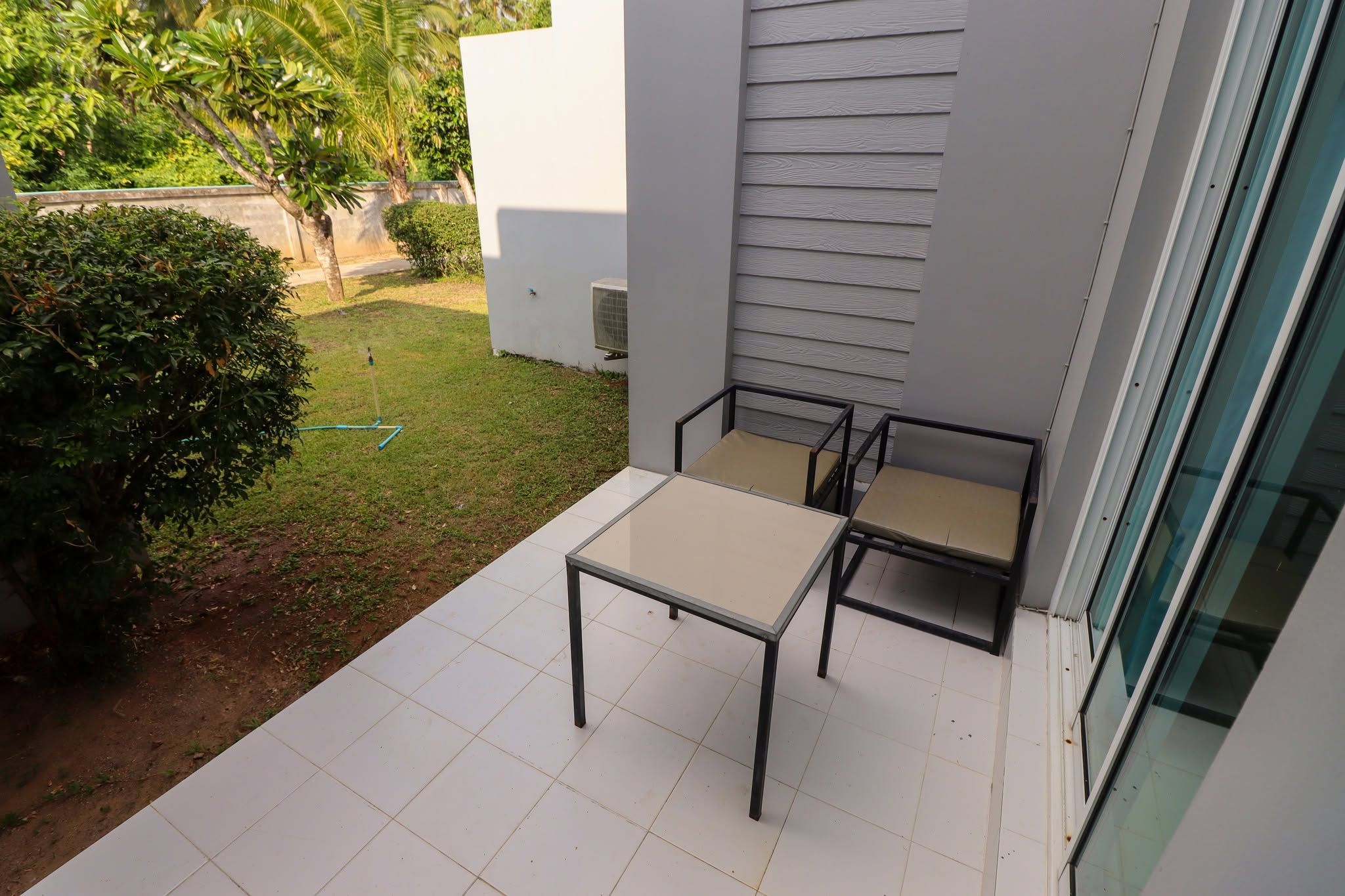 A balcony with a table and two chairs, a small garden area with a hedge, and a building with a white exterior.