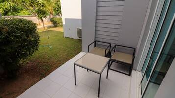 A balcony with a table and two chairs, a small garden area with a hedge, and a building with a white exterior.