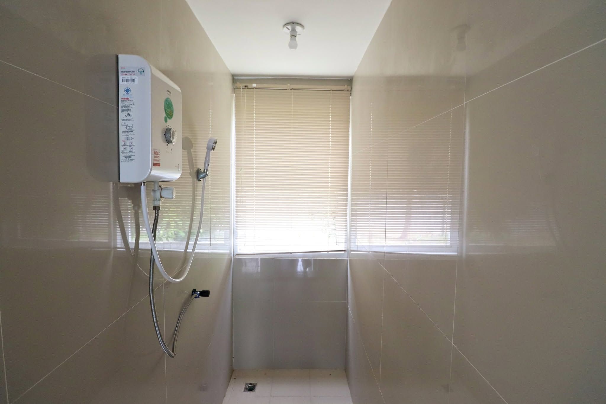 A small, tiled bathroom with a shower unit, a window with blinds, and a ceiling light.