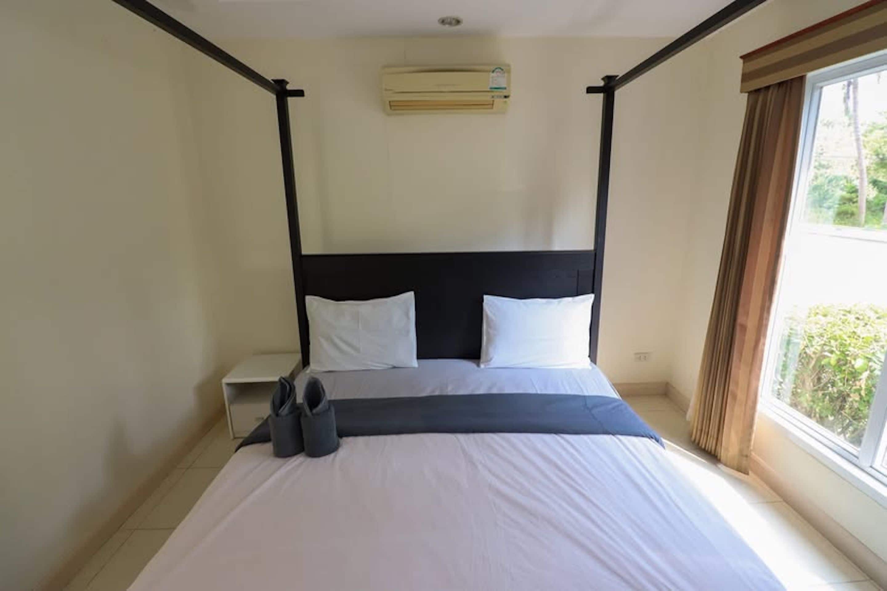 A standard hotel room with a four-poster bed, a bedside table, an air conditioning unit, and a window with curtains.