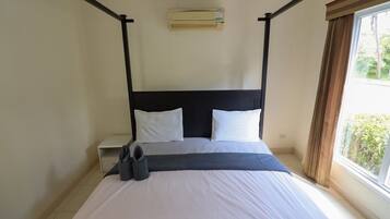A standard hotel room with a four-poster bed, a bedside table, an air conditioning unit, and a window with curtains.