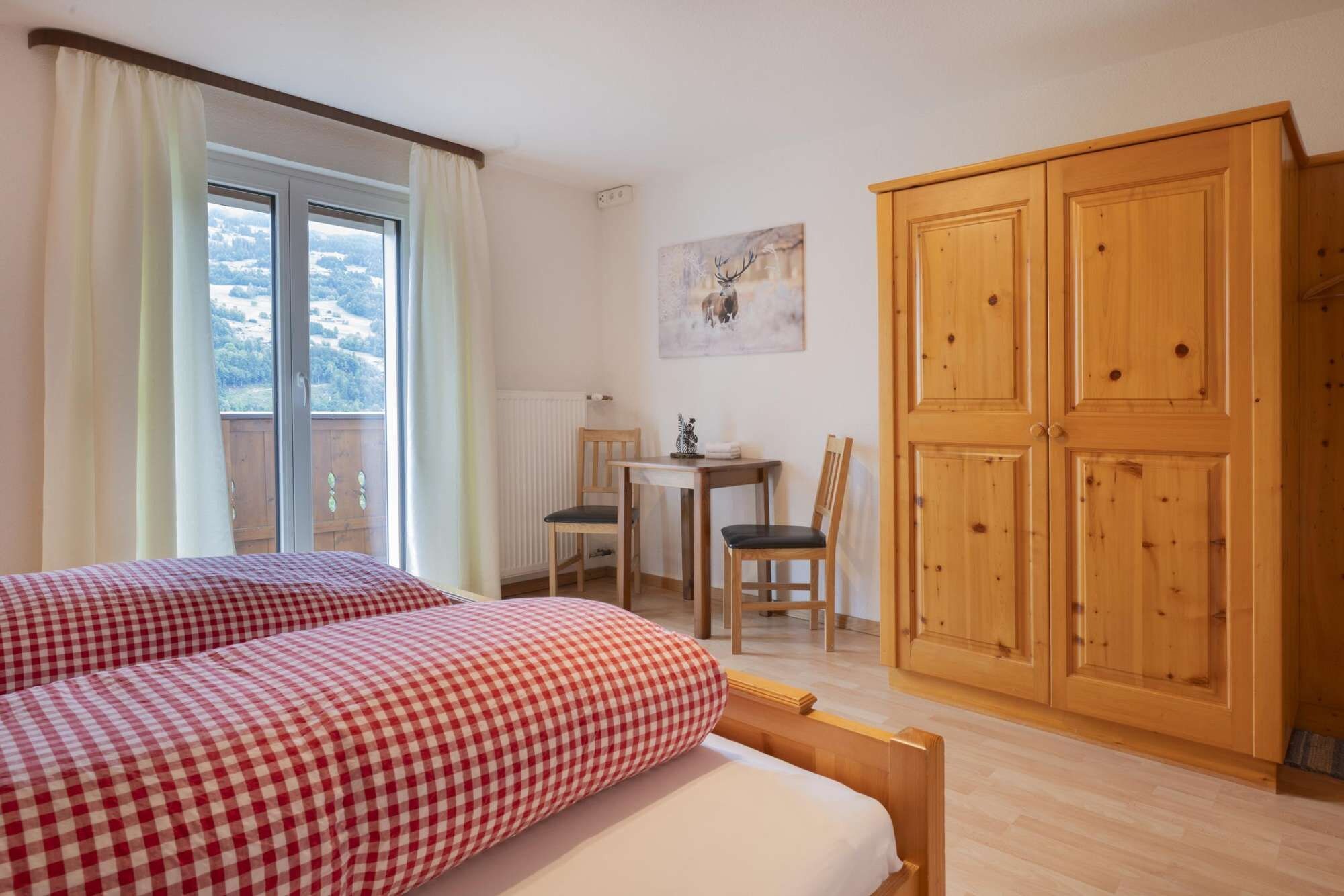 A bedroom with two beds, a wooden wardrobe, a table with chairs, and a painting on the wall.