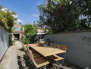 Ein Holztisch mit Stühlen steht draußen, im Hintergrund ist ein Haus zu sehen und eine Baum befindet sich zur Rechten.