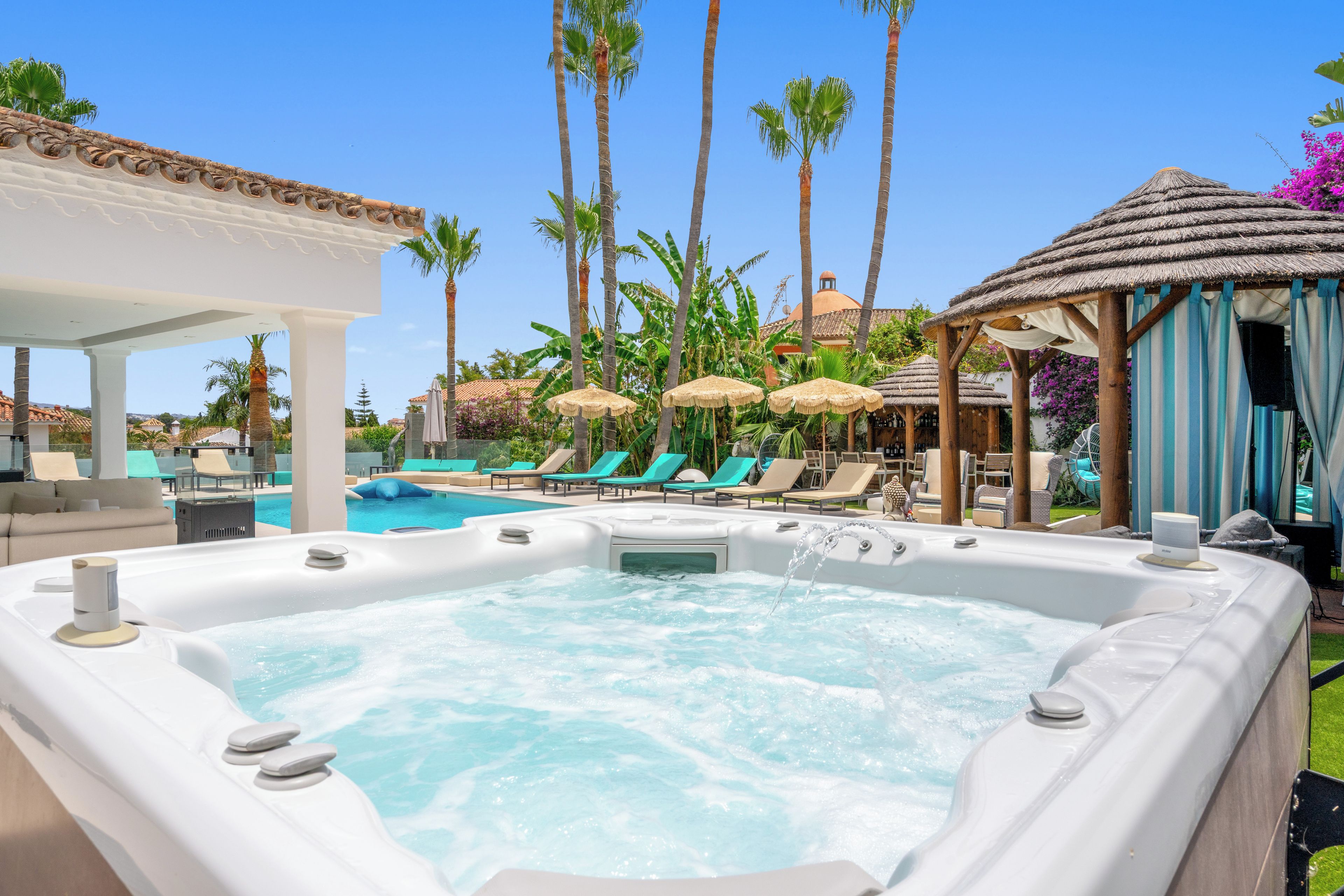 A hot tub surrounded by lounge chairs and umbrellas, with palm trees and a pool in the background.