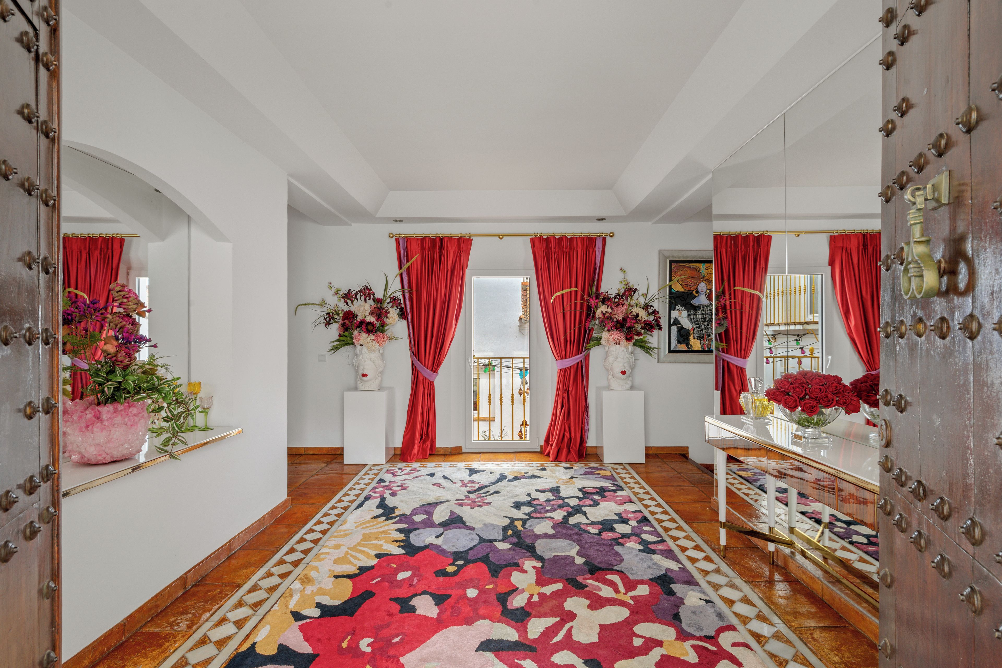 A hallway with a patterned carpet, red curtains, and floral arrangements.