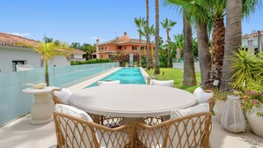 A pool area with a lounge chair and table, surrounded by palm trees and a house with a tiled roof. - Luxury villa in Marbella (Marbella)