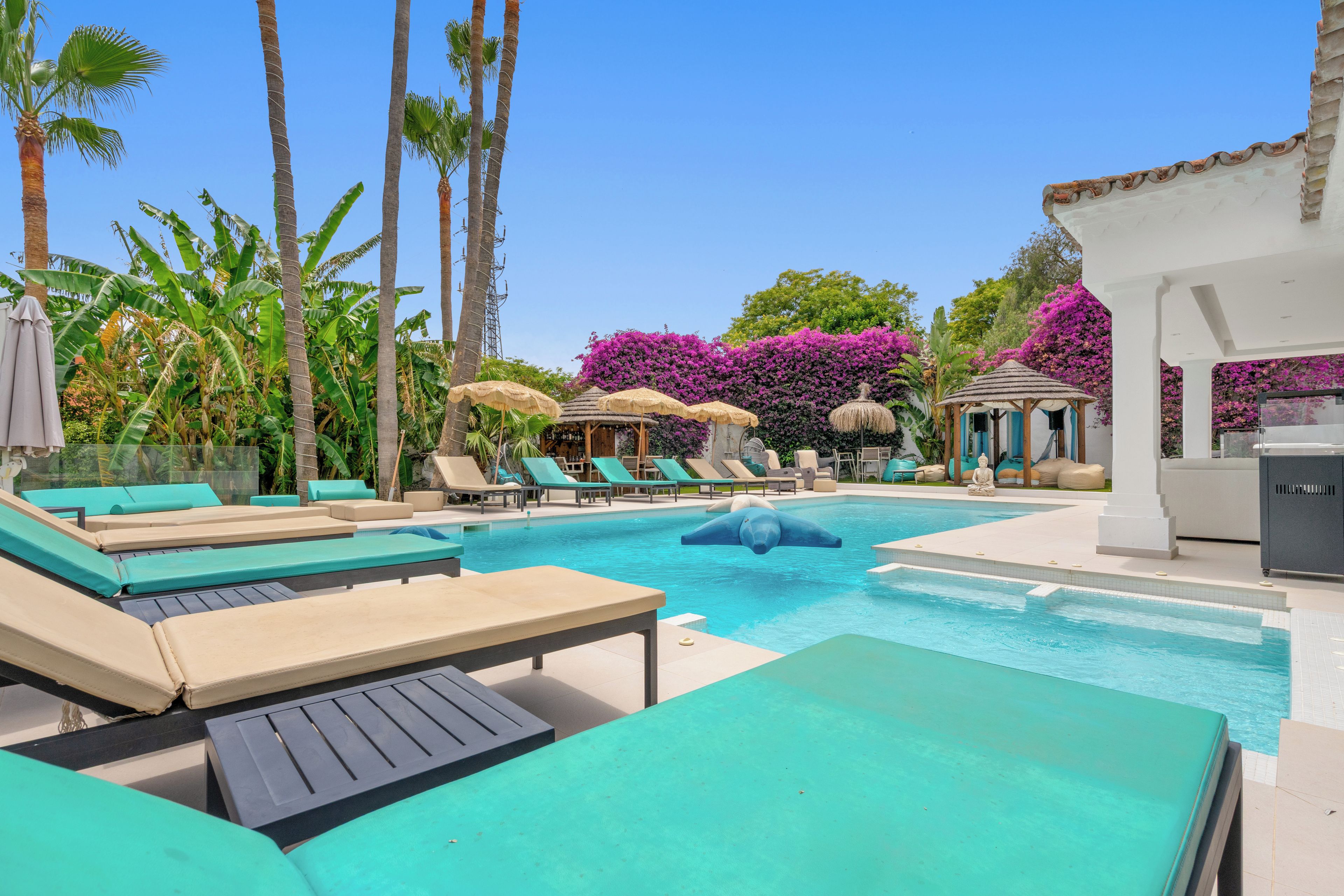 Ein Poolbereich mit Liegestühlen und Sonnenschirmen, umgeben von Palmen und violetten Blumen.