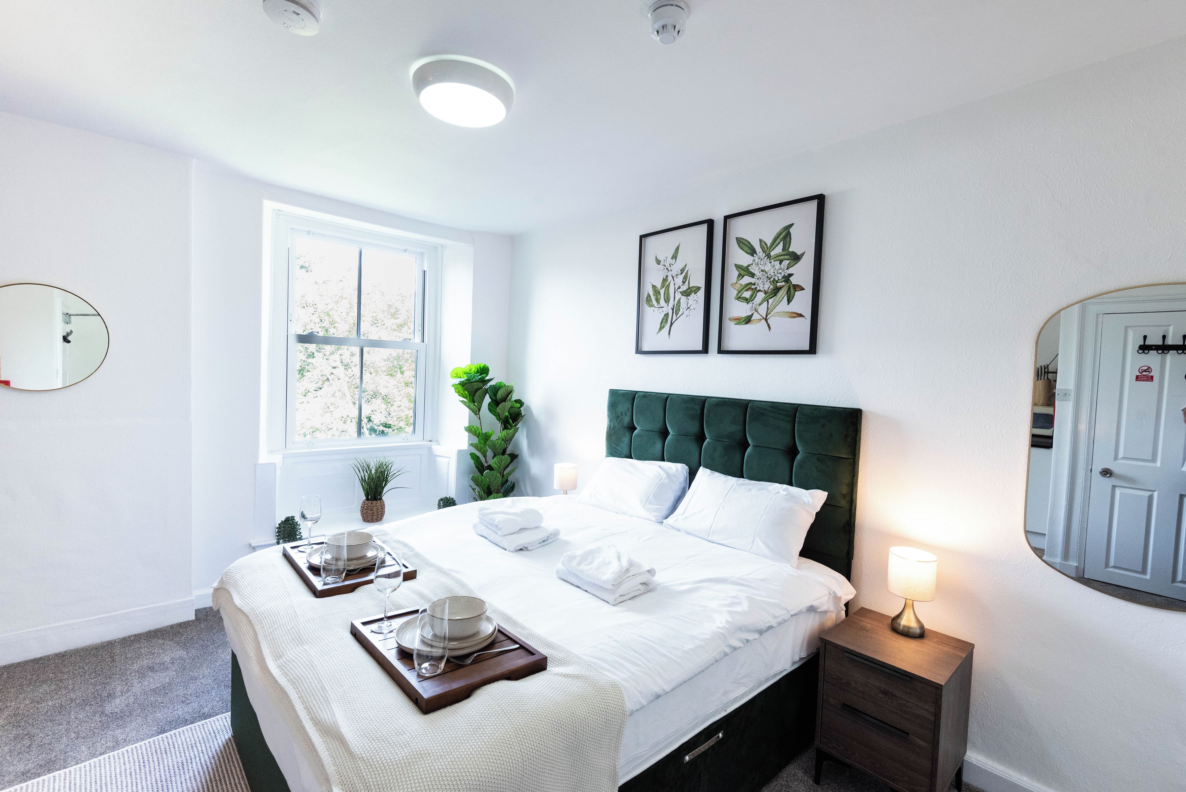 A hotel room with a bed, bedside table, lamp, window, and wall-mounted mirror.