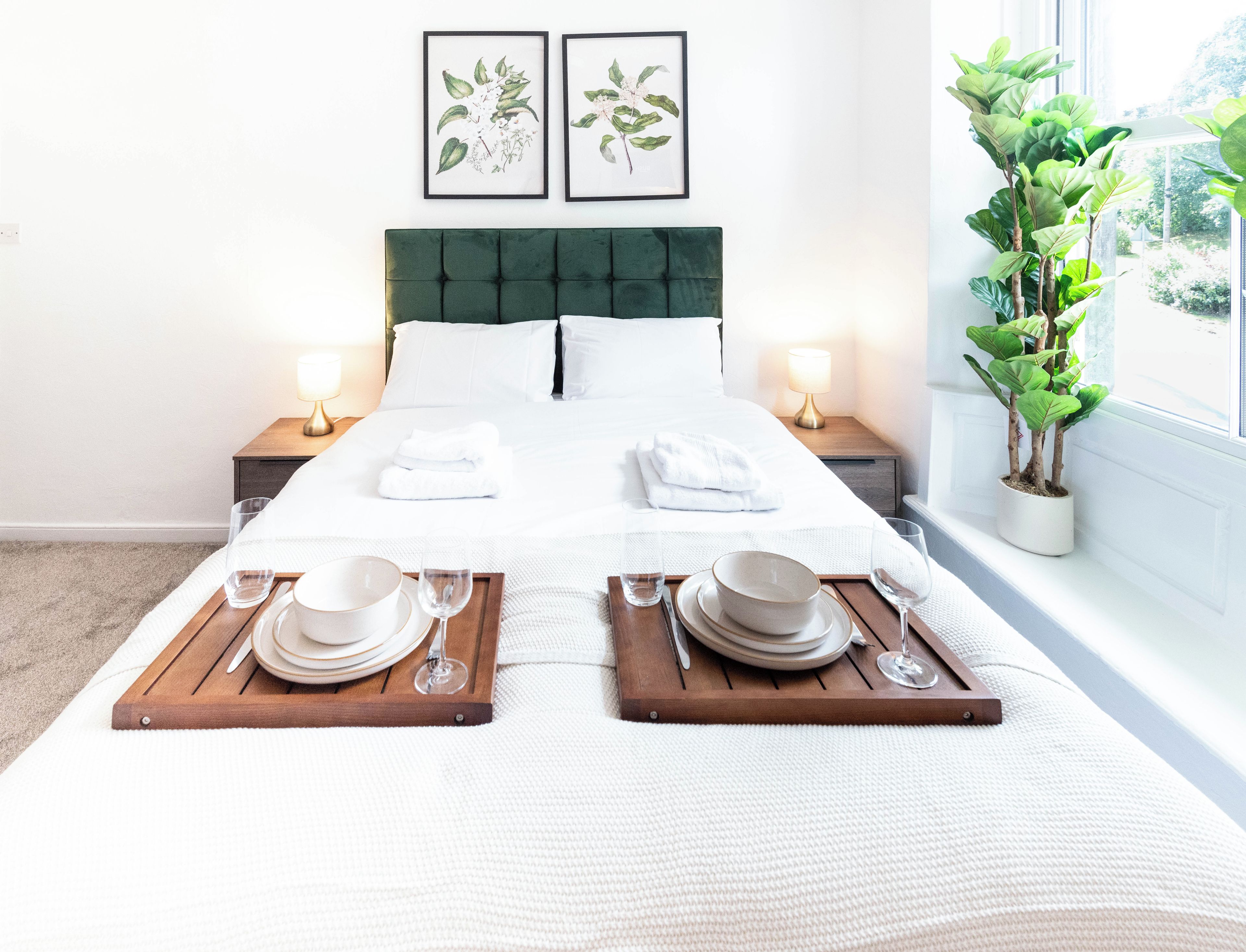 A neatly made bed with white linens, two bedside tables with lamps, a potted plant by the window, and framed botanical prints on the wall.