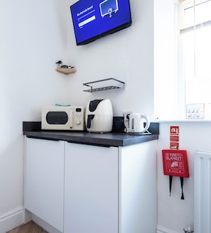 Una kitchenette con microondas, tostadora y hervidor sobre la encimera, un cartel de seguridad contra incendios en la pared y un televisor mostrando un mensaje.