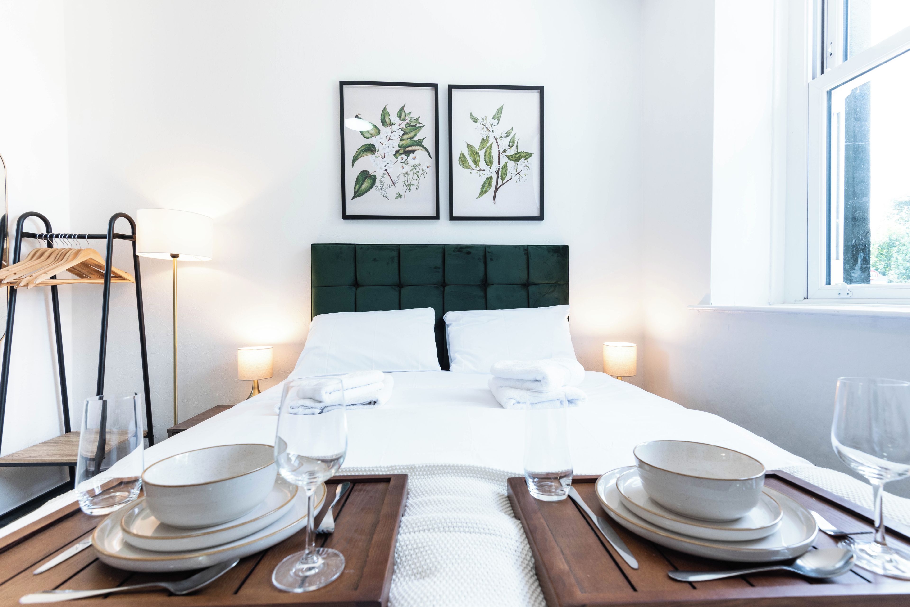 Una cama bien tendida con ropa de cama blanca, un cabecero tapizado en verde y dos impresiones botánicas enmarcadas en la pared.