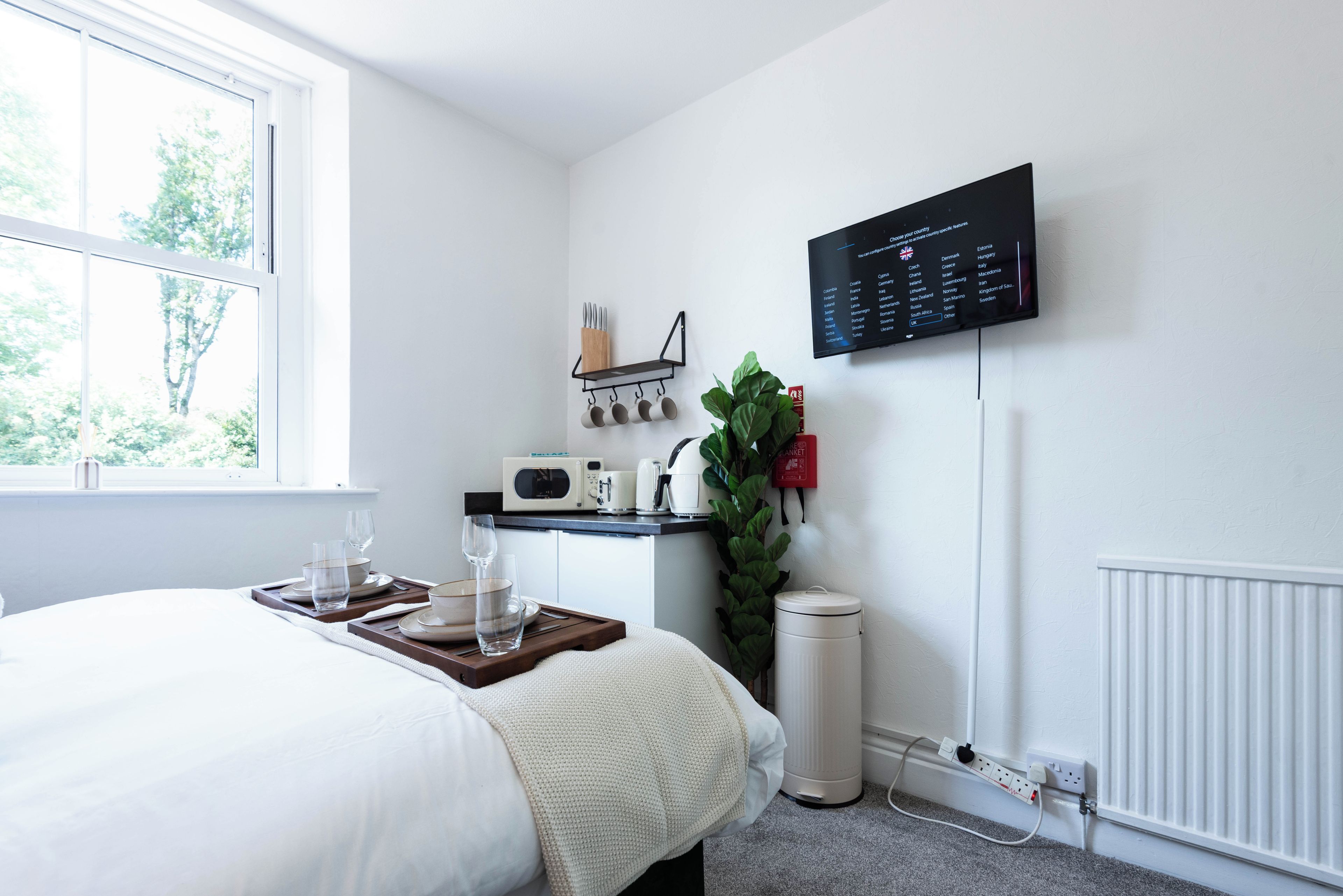 Habitación de hotel con cama, bandeja de desayuno, cafetera, planta y televisor de pared mostrando un menú de selección de canales.