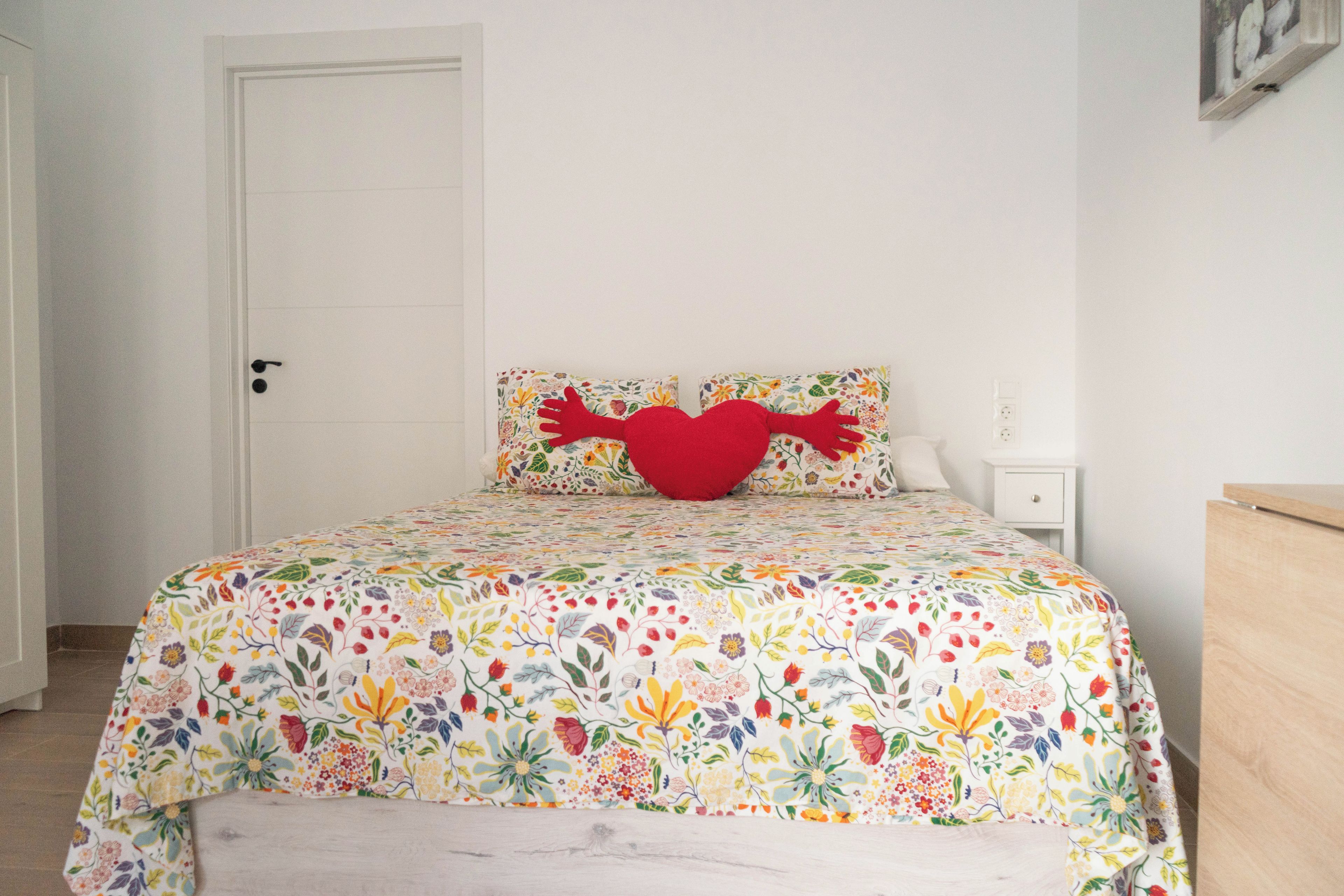Un dormitorio con colcha de estampado floral, un cojín rojo con forma de corazón y una puerta blanca.