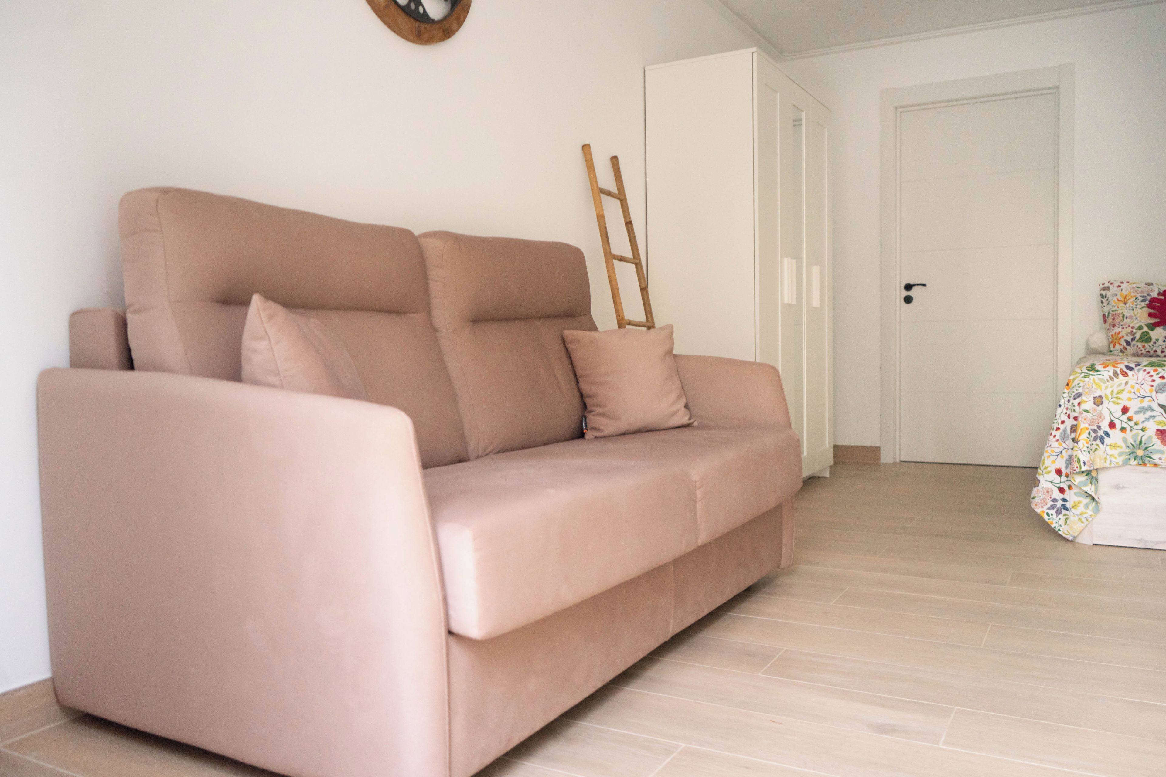 Una habitación con un sofá color beige, una escalera de madera apoyada contra una puerta blanca y una cama con edredón estampado con flores.