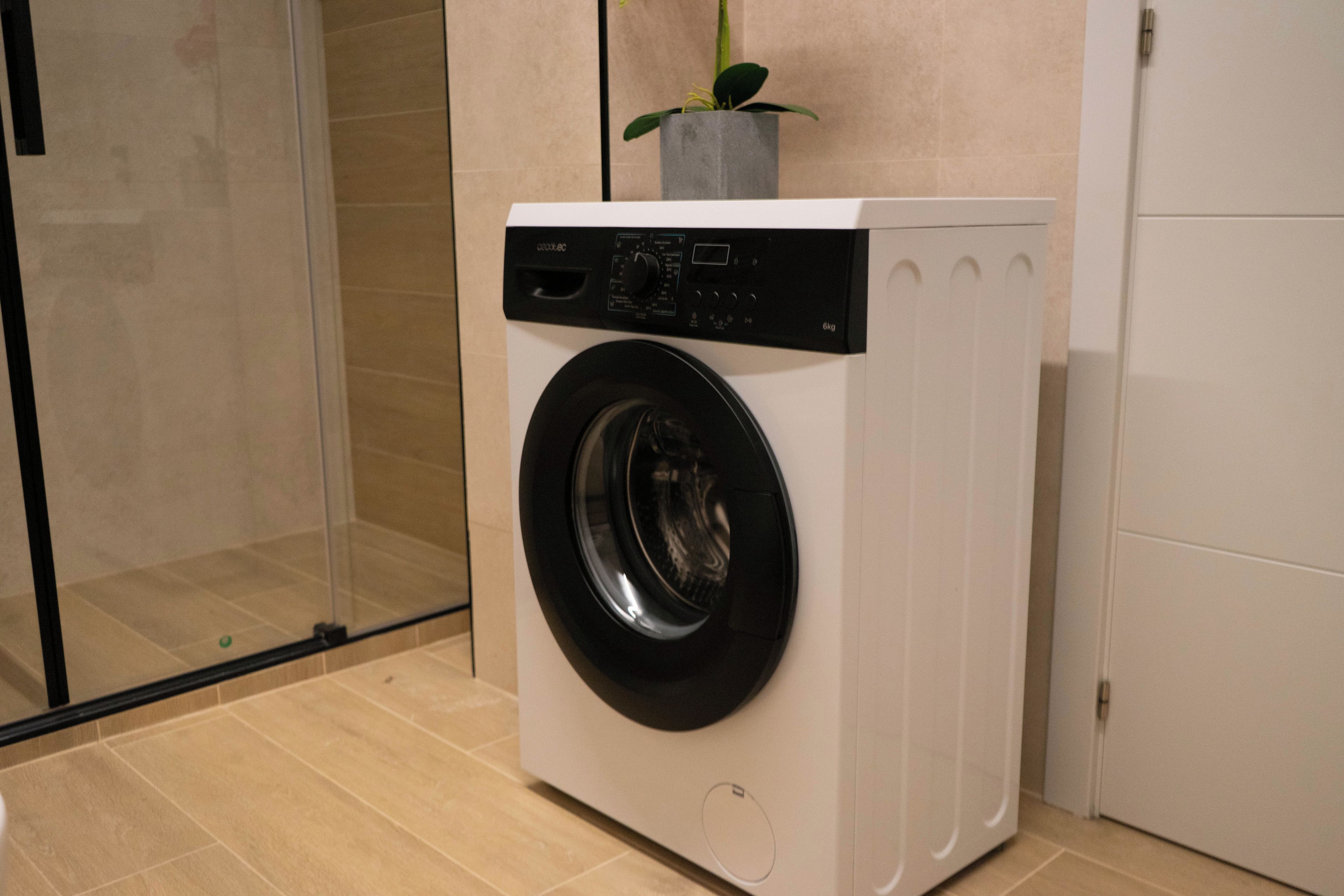 Een wasmachine met een voorlaadopening en bedieningspaneel, een kleine bloempot erbovenop, en een glazen douchedeur op de achtergrond.