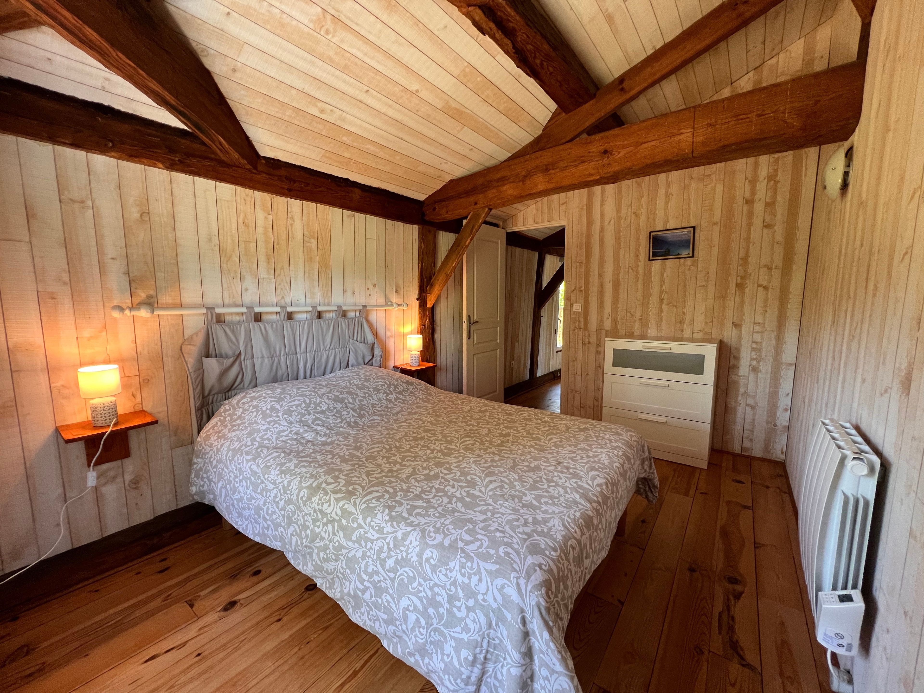 Ein Schlafzimmer mit Holzbalkendecke, einem Bett mit gemustertem Überwurf, Nachttischlampen und einem Heizkörper.
